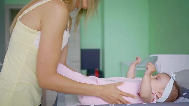 young slim mother is putting her little daughter on a changing table in a room to change a diaper