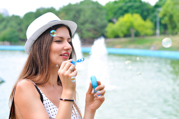 Young cheerful woman in a white hat blowing soap bubbles in a park in the background of a lake