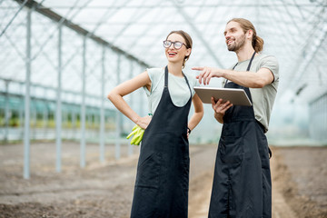 Couple of farmers in uniform working with digital tablet standing in the greenhouse with cultivated...