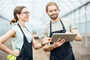Couple of farmers in uniform working with digital tablet standing in the greenhouse with cultivated...