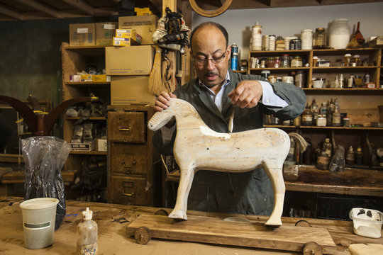 Business owner of antique restoration and wood carving studio at his workbench painting model of wooden horse in New York City, USA