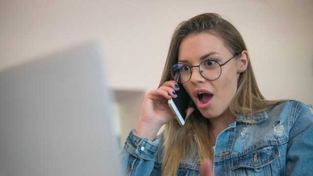 A portrait of young woman who really surprised by speaking on smartphone in front of laptop computer
