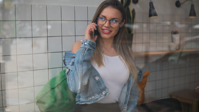 Cute smiling young woman in glasses speaks on the phone through the window