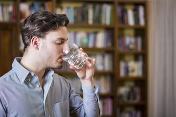 Young man in shirt drinking water from crystal glass on background of bookshelves.