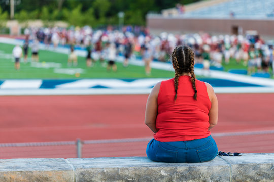 A young lady in red shirt is sitting on a concrete bench beside a football field