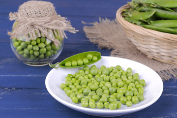 Fresh green peas in a white plate next to peas in a stitch on a blue wooden background