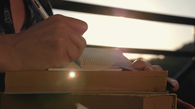 Woman's hand close-up, writing a note or letter to a notebook with a pen on a sunny day at sunset. A stack of scientific books, preparation for exams. Filling out the report