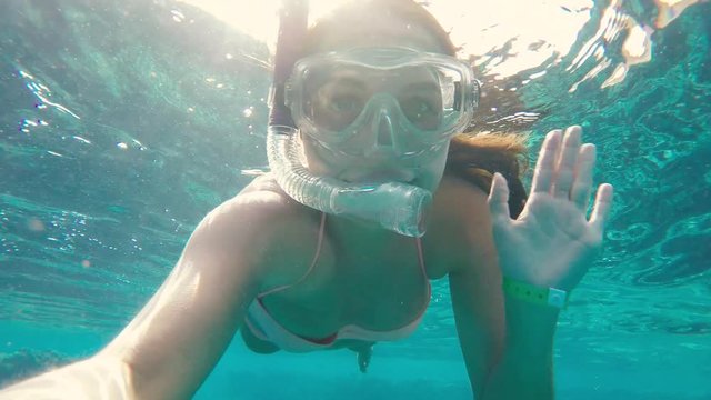 Beautiful young woman in bikini snorkeling with diving mask and waving at the camera while swiming in clear blue water in Red sea. Underwater world with fishes and coral reef. Girl making selfie