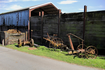 Vintage rusty old farm equipment lying unused on a grass verge in a farmyard, against a large wooden fence with corrugated iron barn on a sunny summer day