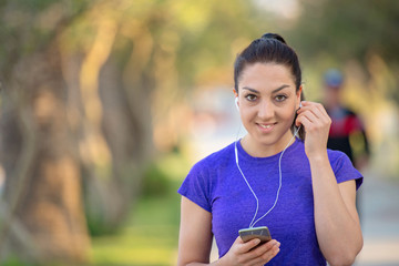 Young woman runner wearing armband and listening to music on earphones. Fit sportswoman taking a break from outdoors training.