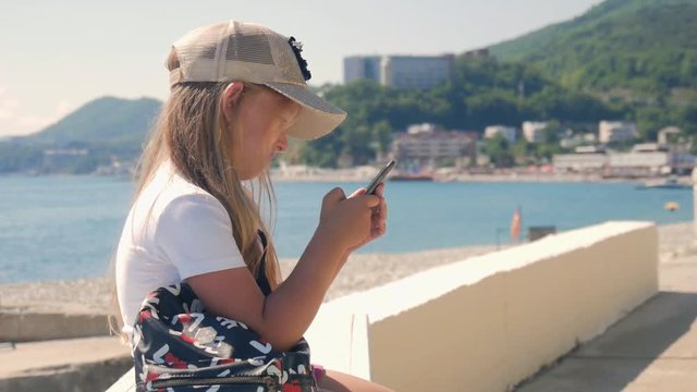 Young girl using a Mobile Phone Sitting on Embankment near the Sea in the Summer. Child controls Smartphone with finger gestures and looks at the social networks.