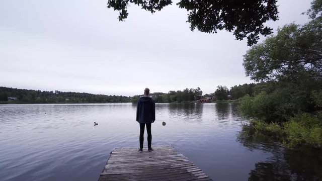 A stylish young man standing on a small pier by a calm lake in Sweden. A smooth gimbal shot moving towards the model. Beautiful outdoor scenery on a summer night