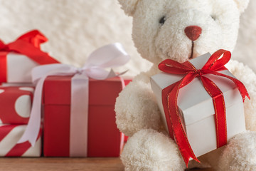 Close-up of teddy bear holding a gift tied with red ribbon on wooden table, white wool background