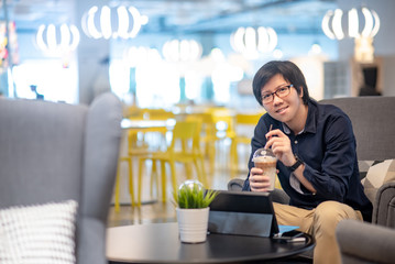 Young Asian happy businessman dressed in casual style drinking coffee working with digital tablet in cafe. Freelance lifestyle or digital nomad concepts