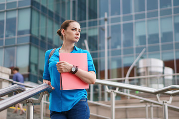 Fototapeta premium Young attractive girl is holding a folder for documents. The manager stands on the steps of the business center and looks thoughtfully sideways.