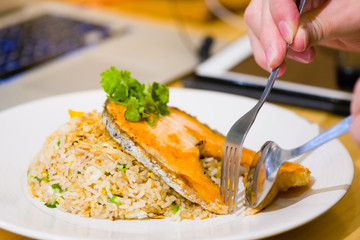 fried sliced salmon with fried rice.man hands use fork and spoon for fusion food.sliced salmon with rice on white plate in restaurant or cafe.man sitting and eating sliced salmon on laptop background