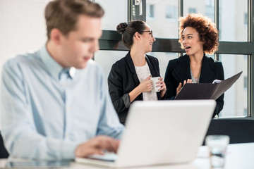 Two young cheerful female employees talking in the office next to their colleague working at desk