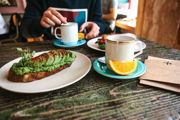 On the table is a useful veggie sandwich or a toast with avocado and a cup next to fresh tea and orange. A man is having breakfast in a cafe.