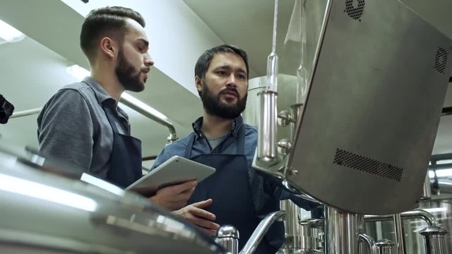 Tilt up shot of bearded Asian worker and his Caucasian colleague using tablet, working with brewing machine at beer plant and discussing something