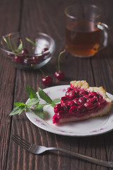 A piece of cherry pie and a sprig of mint lies on a white plate. Behind lie cherry berries and there is a transparent glass cup with tea. All the items lie on an old wooden table.