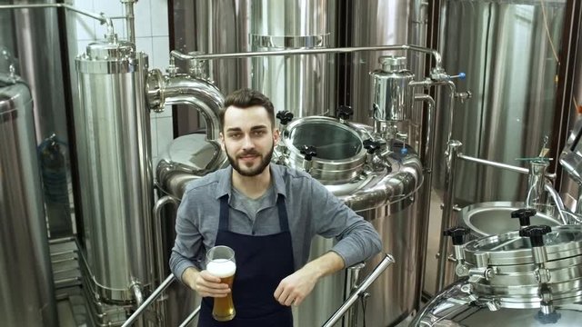 Young handsome man in apron standing at brewing machine at brewhouse, holding glass of freshly brewed craft beer and looking at camera