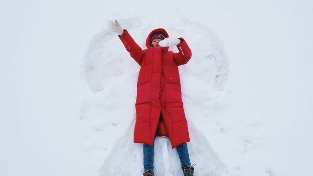 Smiling young woman lies on the snow on a snow angel, waving her hands and sends an air kiss