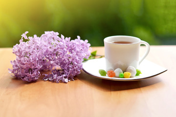 A mug of hot black tea, lilac twigs, a tea rose and sweets (sweets) on a wooden table in the spring at the dacha. Food and drinks.