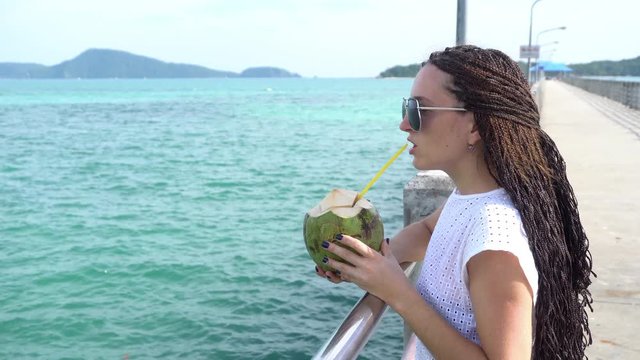 Beautiful girl is drinking coconut water on the beach. The girl wears long zizi braids and sunglasses.