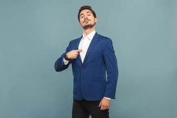 Proud man pointing finger himself. portrait of handsome bearded businessman in blue suit and white shirt, with smart watch. Indoor studio shot, isolated on light blue background