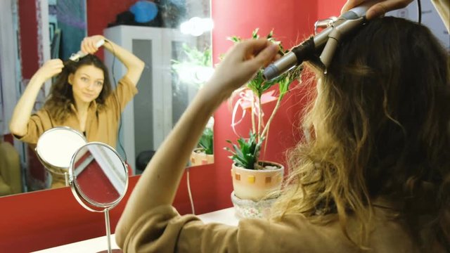 Young beautiful girl looks in the mirror and curls curls on the curling iron in the dressing room, smooth focus