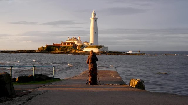 Photographer on causeway takes a photo of St Mary's Lighthouse during golden hour just before sunset.