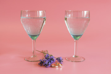 Wedding rings with glasses of wine and lavender flowers on a pink background