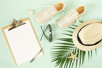 Mock up with clipboard,  women travel clothes straw hat, sneakers espadrill, sunglasses , palm leaf and a pen on pale mint green background. Flat lay, top view.Copy space