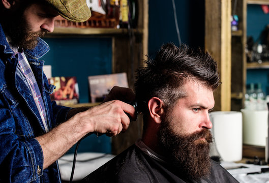 Hipster client getting haircut. Barber with hair clipper works on hairstyle for man with beard, barbershop background. Haircut concept. Barber makes hairstyle using clipper at adult man with beard - Powered by Adobe