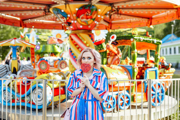 Fototapeta na wymiar Bright summer image. The girl is blonde in a bright striped shirt dress walking in the amusement Park. Portrait of a woman on vacation. She's fooling around and having fun with the carousel.