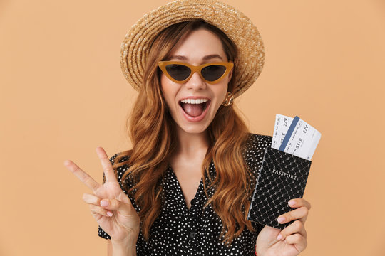 Happy european woman 20s wearing straw hat and sunglasses laughing while holding passport with tickets, isolated over beige background