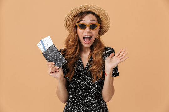 Image of excited brunette woman 20s wearing straw hat and sunglasses screaming while holding passport with travel tickets, isolated over beige background