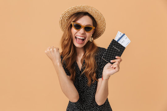 Emotional summer woman 20s wearing straw hat and sunglasses smiling while holding passport with tickets, isolated over beige background