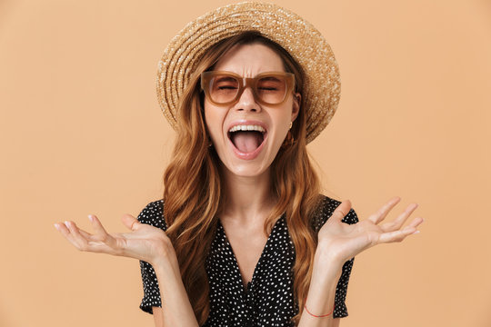 Photo of unhappy capricious woman 20s wearing straw hat and sunglasses throwing arms aside and screaming, isolated over beige background