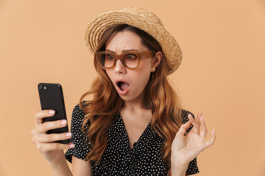Portrait of outraged confused woman wearing straw hat and sunglasses holding and looking at mobile phone, isolated over beige background