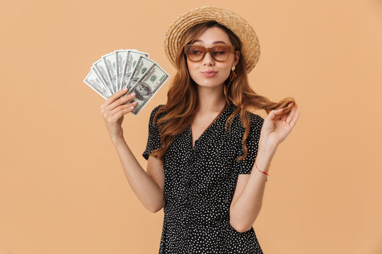 Image of beautiful rich woman 20s wearing straw hat and sunglasses smiling and holding fan of dollar money, isolated over beige background
