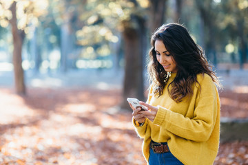 Casual cheerful young woman texting on smartphone outdoor in autumn at city park.