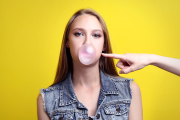 Young woman blowing bubble by chewing gum on yellow background