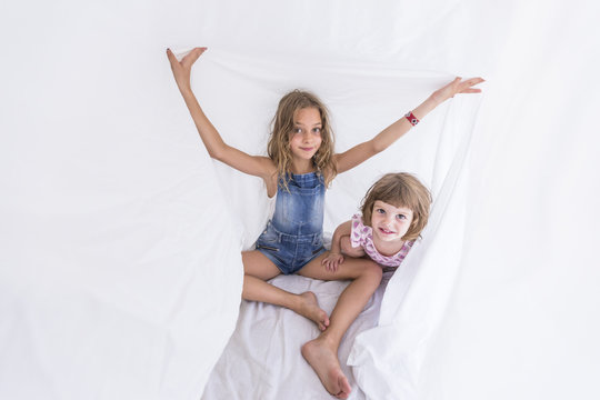 Two Beautiful Sister Kids Playing Under White Sheets On Bed. Fun Indoors. Family Love And Lifestyle