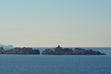 Fototapeta na wymiar A small light house with a red roof is on an island of rocks. The sky is blue with no clouds.