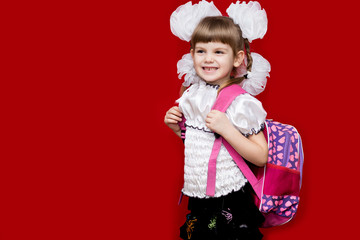 Cute smiling little girl in school uniform and white bows with backpack on red background. Back to school. Education and school concept