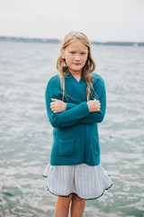 Outdoor portrait of adorable little girl playing next to lake, wearing white dress and green cardigan jacket