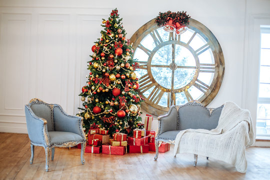 Christmas and New Year decorated white interior room with presents and New year tree with red decor ball. Grey sofa and chair and bid window clock on the wall