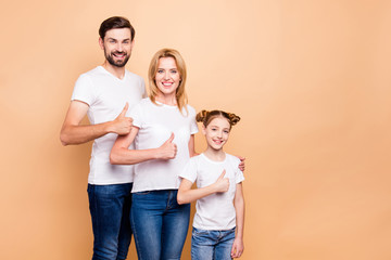 Happy adorable attractive smiling family, bearded father, blonde mother spouses and their little daughter standing straight wearing wtihe t-shirts, blue jeans and showing thumbs up on beige background