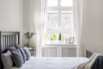 Knit blanket and pillows on bed in white hotel bedroom interior with drapes at window. Real photo